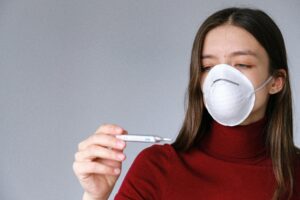 A young woman wearing a mask checks her temperature using a thermometer, highlighting health and safety concerns during a pandemic.